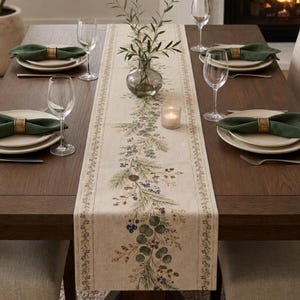 May include: A beige table runner featuring a printed botanical design of pine branches, eucalyptus leaves, and dark berries. The runner is centered on a dark wood table set with white plates, dark green napkins in gold rings, and clear wine glasses.