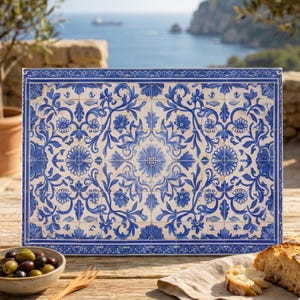 May include: Rectangular ceramic tile with a blue and white floral design. The tile features a border and a central floral motif. The background is a light cream color. The tile is displayed outdoors with a view of the ocean.