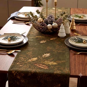 May include: A dark green table runner with a gold botanical print featuring oak leaves and acorns. The table is set with white plates, gold cutlery, a wire bowl filled with pinecones and ornaments, and small white and gold tree figurines.