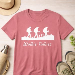 Puede incluir: Camiseta rosa polvoriento con un gráfico blanco de excursionistas y el texto "Walkie Talkies". La camiseta está hecha de un material suave y es adecuada para entusiastas del aire libre. Botas de senderismo y un sombrero son visibles en el fondo.