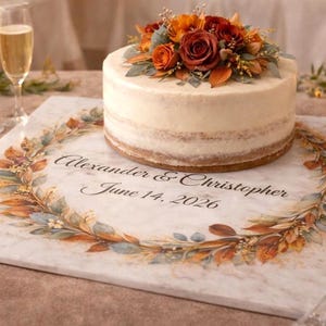 May include: A round, semi-naked cake topped with orange and red roses and greenery, resting on a white marble board printed with a floral wreath and the text Alexander & Christopher June 14, 2026, next to a glass of champagne.