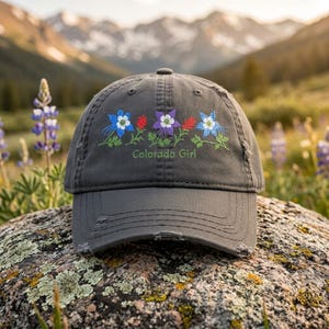 Könnte beinhalten: Eine anthrazitfarbene Baseballkappe mit Used-Optik, verziert mit gestickten blauen und violetten Akeleiblüten, roten Akzenten und grünen Stielen. Der Schriftzug Colorado Girl ist in grünem Garn unter dem Blumenmotiv eingestickt.