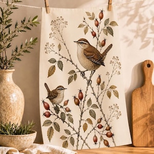 Könnte beinhalten: Ein cremefarbenes Küchentuch mit einer botanischen Illustration von zwei braunen Zaunkönigen, die auf dornigen Zweigen mit grünen Blättern und kleinen roten Hagebutten sitzen. Das Motiv ist in einem detaillierten, natürlichen Stil gedruckt.