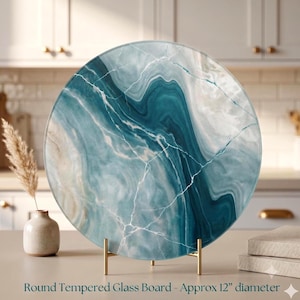 May include: A round tempered glass board with a diameter of 12 inches, featuring a swirling marble-like pattern in shades of teal, blue, and white with thin white veins. The board is supported by a gold-toned metal stand on a flat surface.