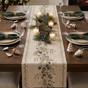 May include: A beige table runner featuring a printed botanical pattern of pine branches and berries, centered with a garland of real pine needles, pinecones, and blue berries, accented by four lit white pillar candles in glass holders.