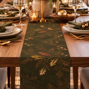 May include: A dark green table runner featuring a printed pattern of golden and bronze-toned autumn leaves, berries, and delicate branches. The runner is centered on a wooden dining table set with white plates, wine glasses, and gold cutlery.