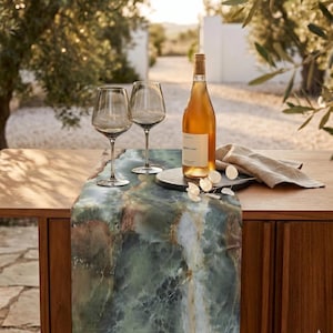 May include: A table runner featuring a green and brown marble-like pattern, two clear glass wine glasses, a bottle of white wine with a label, a round black stone coaster, and a folded beige linen napkin arranged on a wooden table surface.