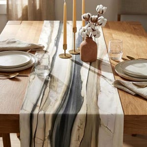 May include: A long table runner featuring a marbled pattern in shades of cream, beige, and charcoal gray. The runner is laid across a wooden table with two gold candle holders, a terracotta vase with cotton stems, and place settings.