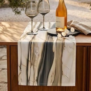 May include: A table runner featuring a marbled pattern in shades of cream, beige, and charcoal gray. The runner is draped over a wooden surface, topped with two clear glass wine glasses, a bottle of white wine, and a beige linen napkin.