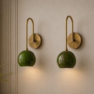 May include: A pair of wall sconces featuring a brushed brass finish on the circular wall mount and the curved arm, paired with a glossy, spherical dark green glass shade that directs light downward from the bottom of the globe.