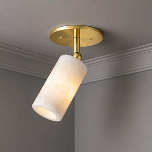 May include: A modern ceiling light fixture with a cylindrical, off-white shade. The light is mounted on a brass-colored base and stem. The fixture is angled, and the light is illuminated, creating a warm glow. The fixture is mounted on a gray ceiling.