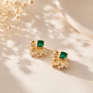 May include: A pair of gold stud earrings featuring a fan-shaped design with a textured finish. Each earring is centered with a square-cut green gemstone and accented with small, clear round-cut stones along the outer edge of the fan shape.