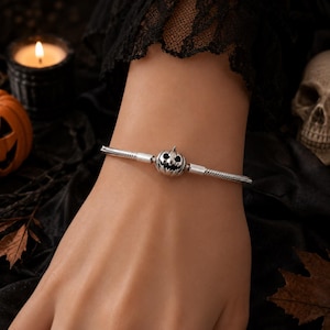 Könnte beinhalten: Ein silberfarbenes Schlangenkettenarmband mit einem zentralen Anhänger in Form eines geschnitzten Jack-o'-Lantern-Kürbisses, der dunkle, vertiefte Details für Augen und Mund aufweist und mit einem einfachen Verschluss am Handgelenk befestigt ist.