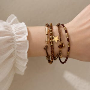 May include: A set of four layered bracelets featuring brown and gold-toned beads, a central gold-colored star charm, and clear faceted glass accents. The bracelets are constructed with a mix of cord and string, showcasing a variety of textures.