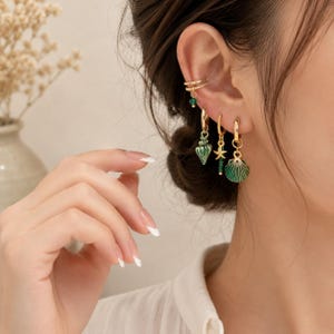 Puede incluir: Un ear cuff dorado con un pequeño abalorio de cuenta verde y tres pendientes de aro dorados con colgantes. Los colgantes incluyen una concha marina verde texturizada, una pequeña estrella de mar dorada con una cuenta verde y una concha.