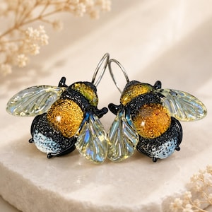 May include: A pair of bee-shaped earrings featuring textured black and amber glass bodies with iridescent, translucent wings. The bee figures are attached to silver-toned metal ear wires, showcasing detailed craftsmanship with a mix of opaque and shimmering glass elements.