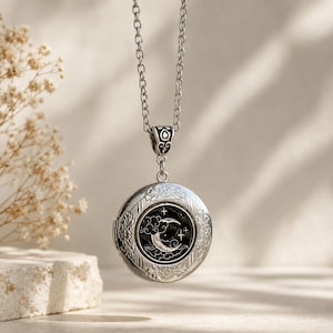 Puede incluir: Un colgante de medallón redondo de plata suspendido de una cadena de plata a juego. El medallón presenta un borde floral intrincado y una incrustación circular central que representa una luna creciente, nubes y estrellas sobre un fondo negro.