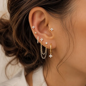 Puede incluir: Una colección de pendientes dorados con varios diseños, incluyendo pequeños broches en forma de flor con piedras transparentes, un broche de estrella, un aro fino, un pendiente conector de doble cadena y un aro con un dije de flor colgante.