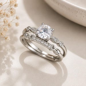 Könnte beinhalten: Ein silberfarbenes Ring-Set bestehend aus zwei Ringen. Der Hauptring hat einen großen, runden, klaren Stein in einer Vier-Krappen-Fassung, flankiert von kleineren Steinen. Der zweite Ring ist ein gerades Design mit einer Reihe kleiner klarer Steine.