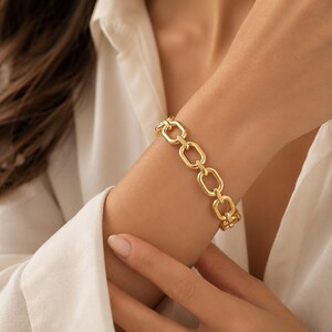 May include: A gold-toned chain bracelet featuring a series of uniform, elongated oval links connected in a repeating pattern. The bracelet has a polished, reflective surface and a secure clasp mechanism, resting on a wrist against a white fabric.