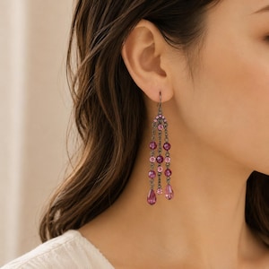 Puede incluir: Un pendiente tipo candelabro con tres cadenas colgantes de metal oscuro, adornado con cristales facetados de color rosa y magenta en varias formas redondas y de lágrima, suspendido de una pequeña pieza decorativa con acentos de cristales rosas.