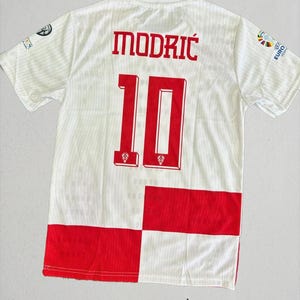 May include: White soccer jersey with red accents, featuring the name "MODRIC" and the number "10" in red. The jersey has a checkered pattern at the bottom and logos on the sleeves. The text "sporysupply" is at the bottom.