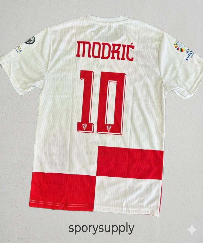 May include: White soccer jersey with red accents, featuring the name "MODRIC" and the number "10" in red. The jersey has a checkered pattern at the bottom and logos on the sleeves. The text "sporysupply" is at the bottom.