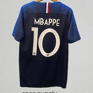 以下が含まれることがあります： 紺色のサッカーユニフォーム。背中には白で「MBAPPE」の名前と「10」の数字がプリントされています。ユニフォームは青い袖と、赤、白、青のストライプが入った襟が特徴です。「sporysupply」の文字が下にあります。