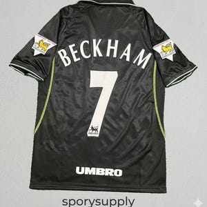 Könnte beinhalten: Schwarzes Fußballtrikot mit dem Namen "BECKHAM" und der Nummer "7" in Weiß. Es hat grüne und weiße Akzente, das Umbro-Logo und "Champions 1999-1999" Patches auf den Ärmeln. Der Text "sporysupply" befindet sich unten.