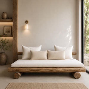 Op de afbeelding: Een rustieke daybed met een houten frame en een wit kussen. De daybed heeft vier kussens, twee grote witte kussens en twee kleinere beige kussens. De daybed staat in een neutrale kamer met een vloerkleed en een houten plank.