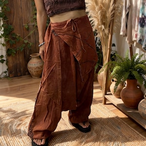 May include: A woman wearing a brown halter top with script and rust-colored wrap pants. The pants have a drawstring waist and button closure. A wooden rack displays various long-sleeved shirts. Decorative vases and plants are in the background.