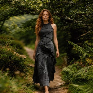 Puede incluir: Una mujer con un vestido halter gris oscuro con un diseño desgastado y en capas camina por un sendero en un bosque. El vestido tiene un cuello alto y una falda asimétrica. El fondo presenta un exuberante follaje verde y un entorno natural.