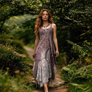 May include: A woman in a long, flowing, sleeveless dress in shades of purple and white, with floral embroidery and lace details. The dress has a layered design and a bohemian style. She is wearing a necklace and walking on a path in a forest.