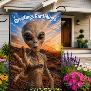 May include: A rectangular garden flag featuring a brown alien with large black eyes reaching forward against a desert landscape at sunset. The text Greetings Earthlings is printed in blue at the top. The flag is displayed on a black metal stand.