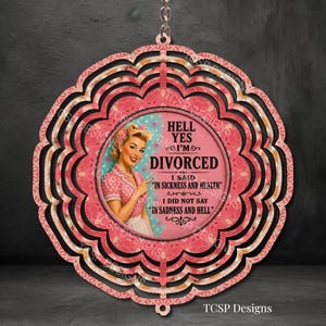 May include: A pink and black decorative hanging sign with a vintage-style image of a woman. The sign reads "Hell Yes I'm Divorced" with additional text. The sign has a scalloped edge and a key ring.