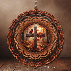 May include: A circular wind spinner featuring a layered, scalloped edge design in shades of brown and orange. The center displays a printed image of a wooden cross in an autumn forest with a red cardinal perched on it and the text TCSP Designs.