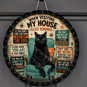 May include: A round decorative wall sign featuring a black cat sitting on a teal armchair. The text reads: WHEN VISITING MY HOUSE PLEASE REMEMBER IF YOU DON'T LIKE CAT HAIR STAY OFF THE SOFA, I LIVE HERE YOU ARE THE GUEST, YOU WILL BE SNIFFED & POSSIBLY LICKED, TO YOU I AM THE CAT BUT HERE I AM THE BABY, THEY LIKE ME MORE THAN THEY LIKE MOST HUMANS, THEY LOVE ME THEY'RE ONLY FRIENDS WITH YOU.