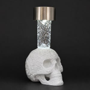 May include: A white skull-shaped base with intricate embossed floral patterns, topped with a clear, faceted glass cylindrical neck and a metallic silver cap. The object features a bright light emitting from the center of the glass section.