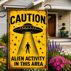May include: A rectangular yellow garden flag with a distressed finish featuring the black text CAUTION at the top and ALIEN ACTIVITY IN THIS AREA at the bottom. A black graphic of a UFO beaming light down on a human silhouette is centered.