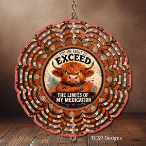 May include: A circular hanging wind spinner featuring a central illustration of a brown Highland cow with crossed arms. The text reads YOU ARE ABOUT TO EXCEED THE LIMITS OF MY MEDICATION. The outer edges have a scalloped, layered floral pattern.