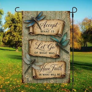 May include: A garden flag with a wooden background and three scrolls with text and dragonfly illustrations. The scrolls read "Accept What Is", "Let Go of What Was", and "Have Faith in What Will Be". The flag is displayed outdoors.
