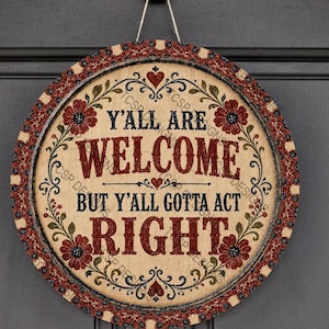 May include: A round wooden sign with a beige background and a dark red floral border. The text reads Y'ALL ARE WELCOME BUT Y'ALL GOTTA ACT RIGHT in dark red and black lettering. The sign features small red hearts and green vine accents.