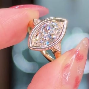 Può includere: Un anello d'oro con un diamante taglio marquise. Il diamante è incastonato in una montatura a castone con piccoli diamanti di accento su ogni lato. L'anello è tenuto da un dito. Il testo "THE DIAMOND EDIT" è nell'angolo in alto a sinistra.