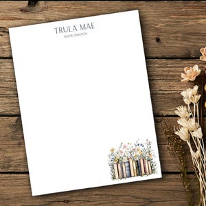 Puede incluir: Papelería blanca con el texto "TRULA MAE BOOK DRAGON" en la parte superior. Una ilustración de acuarela de libros y flores está en la esquina inferior derecha. La papelería está sobre una superficie de madera.