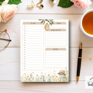 Puede incluir: Bloc de notas beige y blanco con secciones para "To Do", "Date", "Important" y "Notes". El bloc de notas presenta un diseño de abeja y floral. Una taza de té, un bolígrafo y gafas están sobre una superficie de madera blanca.