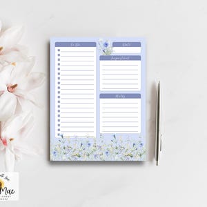 Puede incluir: Un bloc de notas con temática floral con secciones para 'To Do', 'Date', 'Important' y 'Notes'. El bloc de notas tiene un fondo azul claro con un diseño floral en la parte inferior. Un bolígrafo plateado descansa junto al bloc de notas. El bloc de notas está etiquetado 'Trula Mac Stationery & More'.