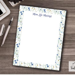 Puede incluir: Una hoja de papelería floral con el texto "Mom Life Musings" en la parte superior. El borde presenta flores azules y blancas y hojas verdes. Un bolígrafo, una taza de café y un ordenador portátil están sobre la superficie de madera.