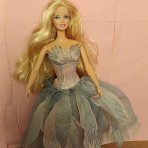 以下が含まれることがあります： 青い目をしたブロンドの人形が、淡いピンクのボディスと、金色のグリッターが施された半透明のグレーブルーの葉の形をした生地のパネルを重ねたスカートが特徴の、ストラップレスのドレスを着用しています。
