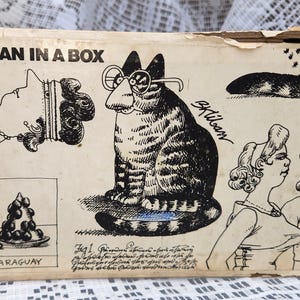 Könnte beinhalten: Ein rechteckiger Pappkarton mit der Aufschrift KLIBAN IN A BOX, der schwarze Tuschezeichnungen einer Katze mit Brille, einer Person mit Halskette, einer mit PARAGUAY beschrifteten Weintraubenschale und zwei Personen zeigt. Die Oberfläche ist cremefarben.