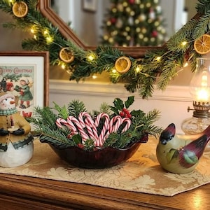 May include: A holiday display featuring a snowman figurine, a dark red bowl filled with pine branches and candy canes, a bird figurine, and a small oil lamp. A framed Christmas print sits behind the snowman, and a key fob rests on a wooden tray.